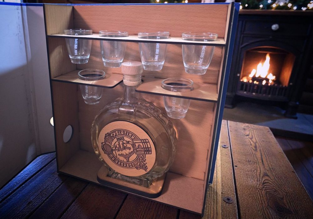 Pijani registrator s drveno ugraviranom bocom i čašicama, postavljen u blagdanskom okruženju s kaminom i borom.