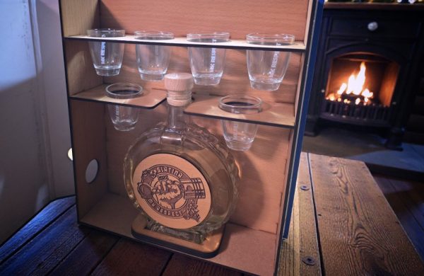 Pijani registrator s drveno ugraviranom bocom i čašicama, postavljen u blagdanskom okruženju s kaminom i borom.